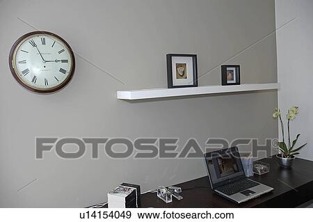 Circular clock on wall above laptop computer on desk in modern grey homeoffice View Large Photo Image Stock Photo - Circular clock on wall above laptop computer on desk in modern grey homeoffice. Fotosearch