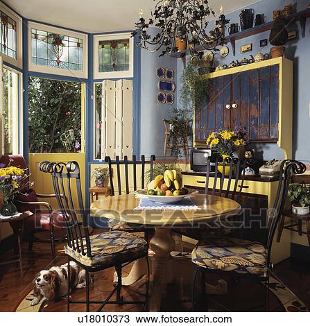 Stock Image - Circular yellow table and iron chairs with colourful cushions in pastel blue dining room with stained glass windows. Fotosearch