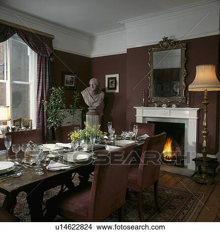Picture - Classical bust in corner of brown dining room with place settings and mirror above lighted fire in fireplace . Fotosearch