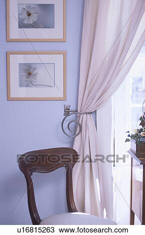 Stock Image - Close-up of antique chair in front of cream curtain with metal tieback beside pictures on pale blue wall. Fotosearch