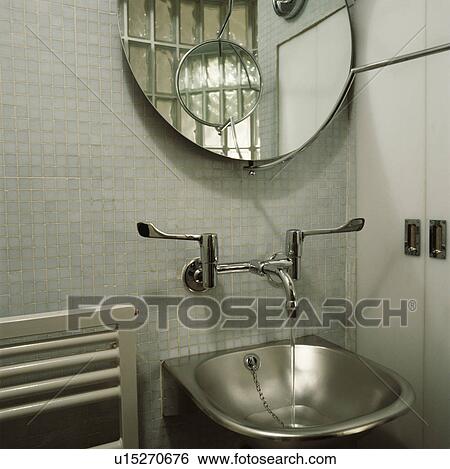 Stock Photograph - close-up of circular mirror above stainless steel wall-mounted taps and basin on mosaic tiled wall. Fotosearch