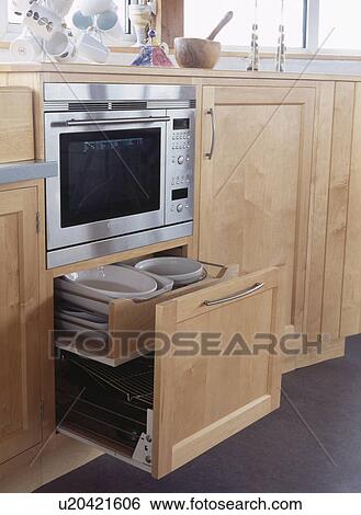 Stock Photograph - Close-up of crockery in open pull-out drawer below oven in pale wood kitchen. Fotosearch