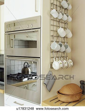 Stock Photo - Close-up of cups stored on metal rack beside double stainless steel oven in modern kitchen. Fotosearch