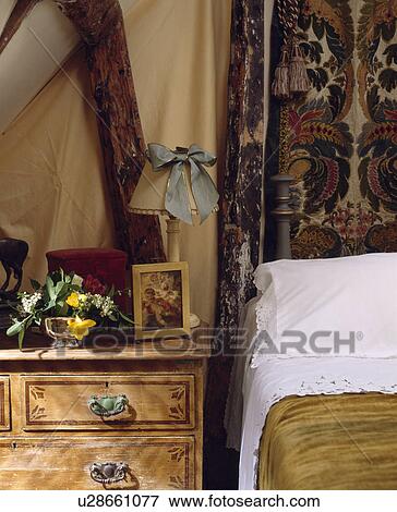 Stock Photo - Close-up of lamp on old chest-of-drawers beside bed with white pillow. Fotosearch