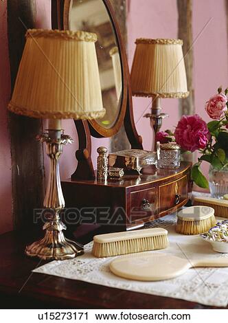 Stock Image - Close-up of metal lamps with cream ruched shades on dressing table with ivory brushes and hand-mirror. Fotosearch