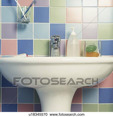 Stock Image - Close-up of toiletries on white basin below multicoloured pastel wall tiles. Fotosearch