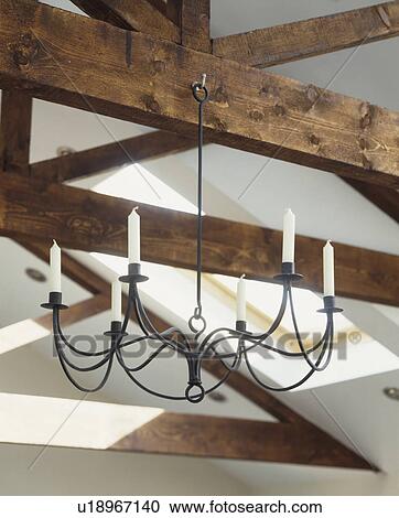 Stock Image - Close-up of white candles on black wrought iron chandelier on wooden ceiling beams. Fotosearch