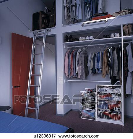 Stock Photo - Clothes on hanging rails in fitted open storage with shelves and metal units beside metal ladder over open door. Fotosearch