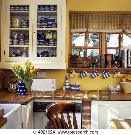Collection of blue+white Cornishware in glass-fronted cream cupboard beside window in yellow cottage kitchen View Large Photo Image Picture - Collection of blue+white Cornishware in glass-fronted cream cupboard beside window in yellow cottage kitchen. Fotosearch