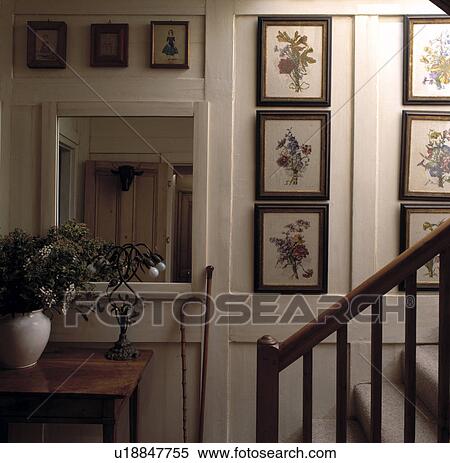 Stock Photography - Collection of floral pictures on cream panelled walls of small cottage hall with square wooden mirror. Fotosearch