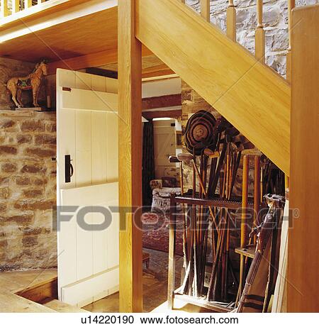 Stock Image - Collection of walking sticks in stand below wooden staircase in cottage hall with cream door open to living room. Fotosearch
