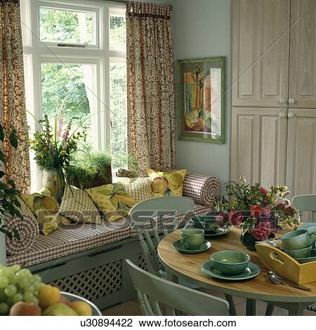 Stock Image - Colourful cushions piled onto checked window seat in pastel green dining room with green crockery and chairs . Fotosearch