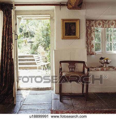 Stock Image - Cottage hall with stone-flagged floor and antique chair beside open door with view of the garden. Fotosearch