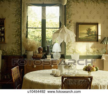 Stock Image - Cotton lampshade on pendant lamp above table with lace cloth in Edwardian style living room with painted floral walls. Fotosearch