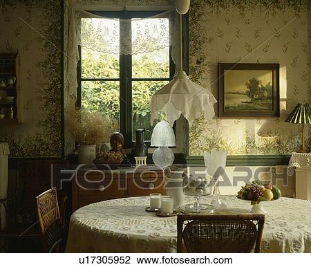 Stock Image - Cotton lampshade on pendant lamp above table with lace cloth in Edwardian style living room with painted floral walls. Fotosearch