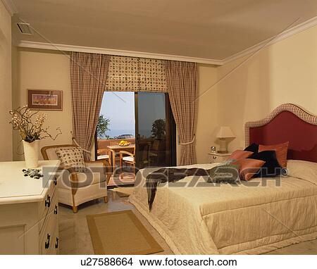 Picture - Cream quilt on bed in neutral bedroom with beige curtains and patterned blind at patio doors to balcony. Fotosearch