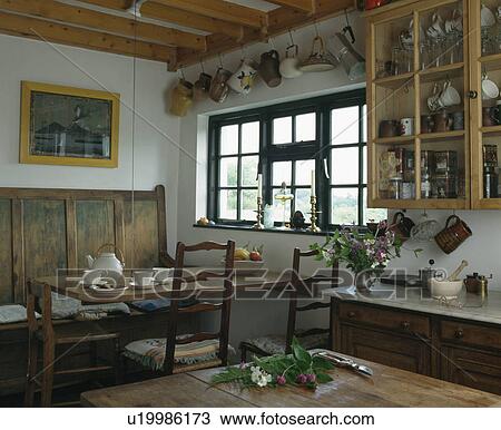 Stock Image - Cream teapot and crockery on wooden table with antique wooden settle in traditional country diningroom. Fotosearch