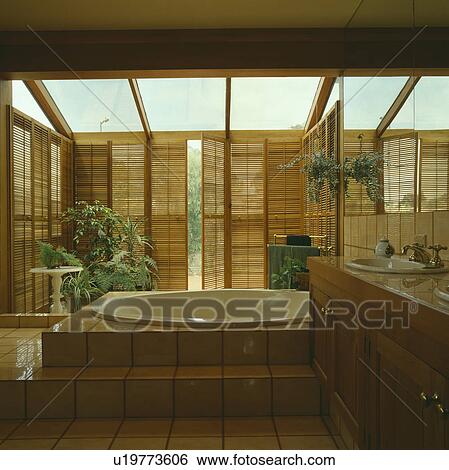 Stock Photograph - Cream tiled steps to white bath in modern bathroom extension with plantation shutters. Fotosearch