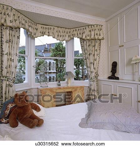 Cream+green floral curtains at bay window in traditional white bedroom with teddybear on bed with white linen View Large Photo Image Stock Image - Cream+green floral curtains at bay window in traditional white bedroom with teddybear on bed with white linen. Fotosearch