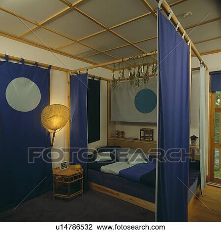 Dark Blue Bedlinen On Fitted Bed With Wooden Poles And Blue Curtains In Eighties Bedroom Stock Image