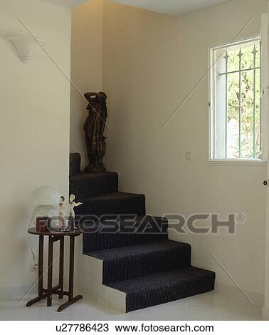 Stock Image - Dark grey carpet on stairs in white hall with ceramic figures on small table. Fotosearch