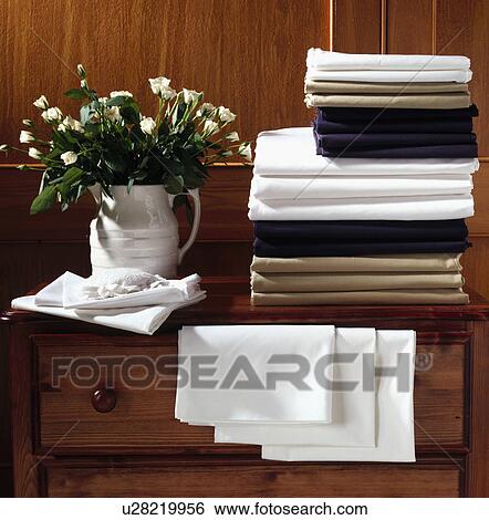 Stock Photograph - Detail of folded bed linen stacked on wooden chest of drawers with white jug of flowers. Fotosearch