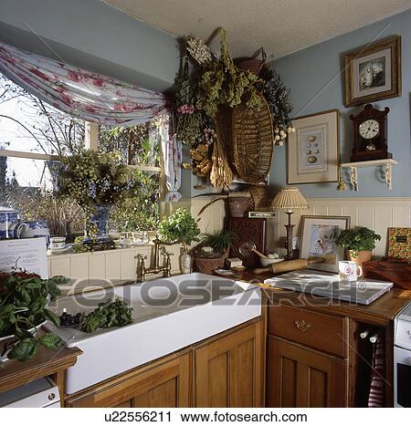Stock Image - Dried flower arrangement in corner of pastel blue kitchen with double white ceramic sink below window. Fotosearch