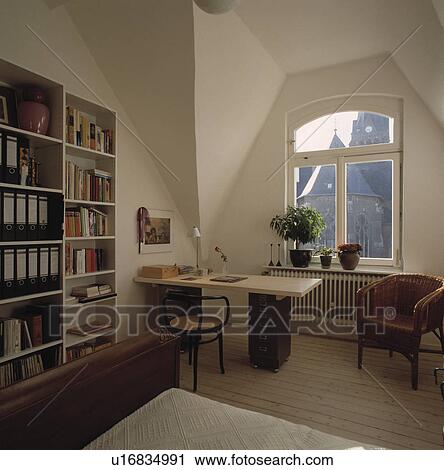 Stock Image - Files and books on white shelves in white attic bedroom with office desk and chair. Fotosearch