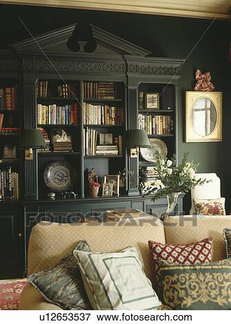 Fitted bookshelves with pediment in dark green living room with cushions piled on cream sofa View Large Photo Image Stock Photo - Fitted bookshelves with pediment in dark green living room with cushions piled on cream sofa. Fotosearch