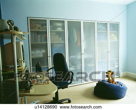 Stock Image - Fitted storage cupboards with opaque glass doors in home office bedroom with computer on desk and beanbag on floor. Fotosearch