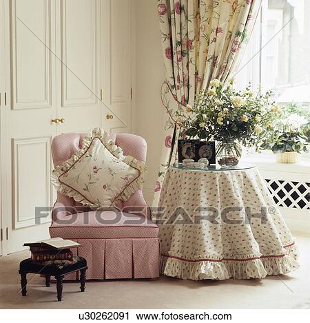 Stock Image - Floral cushion on pink armchair beside circular table with floral cloth in traditional bedroom. Fotosearch