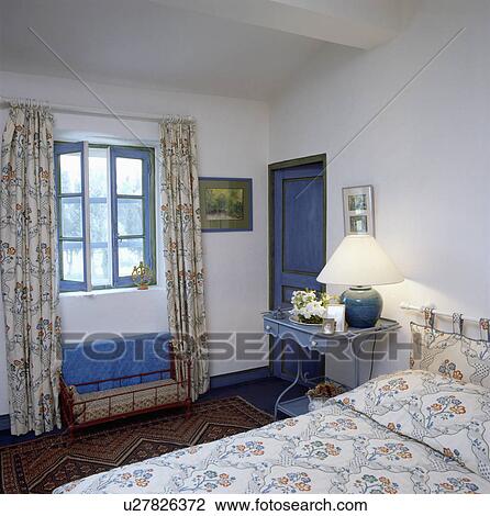 Floral quilt on bed and matching curtains on window in white country bedroom with lighted lamp on blue bedside table View Large Photo Image Stock Image - Floral quilt on bed and matching curtains on window in white country bedroom with lighted lamp on blue bedside table. Fotosearch