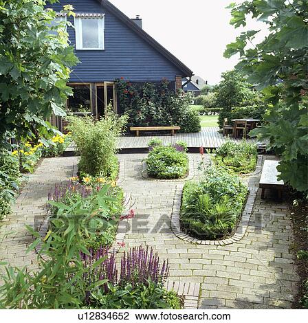 Stock Image - Flower beds in paving in Scandinavian country garden with small blue house. Fotosearch