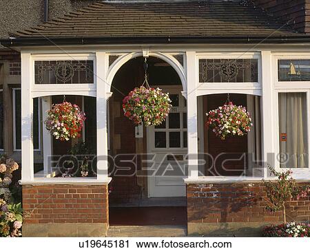 Stock Image - Flowering plants in hanging baskets on glass porch of traditional house. Fotosearch