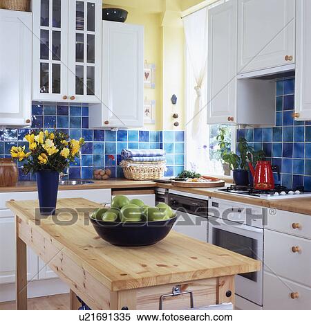 Stock Photography - Flowers and bowl of apples on pale wood table in white kitchen with fitted oven and dishwasher and blue wall tiles. Fotosearch