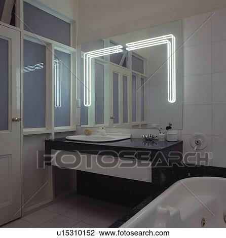 Stock Image - Fluorescent lighting in large mirror above basin in vanity unit in modern white bathroom. Fotosearch