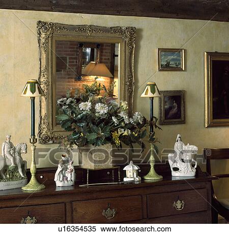 Gilt framed mirror above chest of drawers with collection of Staffordshire figures and lamps on either side of a flower arrangement View Large Photo Image Stock Photography - Gilt framed mirror above chest of drawers with collection of Staffordshire figures and lamps on either side of a flower arrangement. Fotosearch