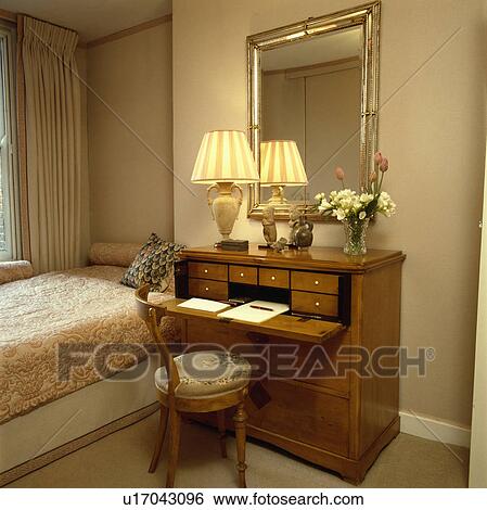 Stock Photograph - Gilt mirror above lighted lamp on antique walnut desk anc chair beside single bed with patterned quilt. Fotosearch