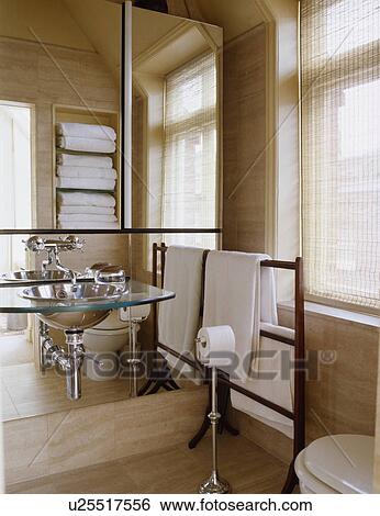 Stock Photograph - Glass and stainless steel basin on mirrored wall in modern bathroom with white towels on wooden towel rail. Fotosearch