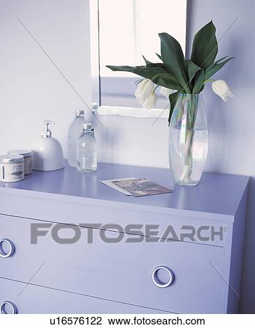 Stock Image - Glass vase of white tulips on top of a lilac painted chest of drawers . Fotosearch