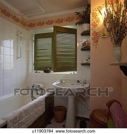 Picture - Green louvre shutters on window in bathroom with white tiled wall above bath and stencilled floral garland on peach walls . Fotosearch