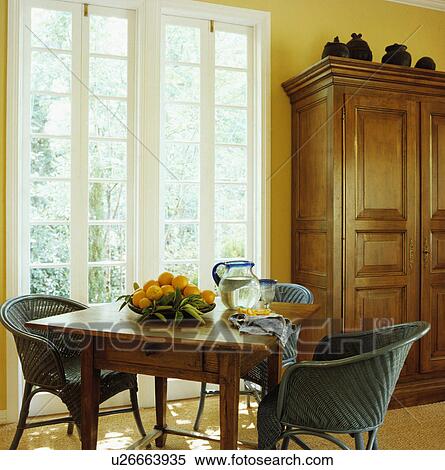 Stock Photography - Green wicker chairs and old pine table in front of French windows in yellow dining room with large cupboard. Fotosearch