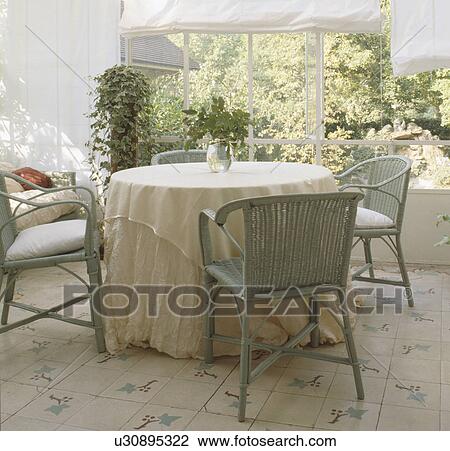 Stock Image - Grey wicker chairs and circular table with cream cloths in conservatory dining room with white cotton blinds. Fotosearch