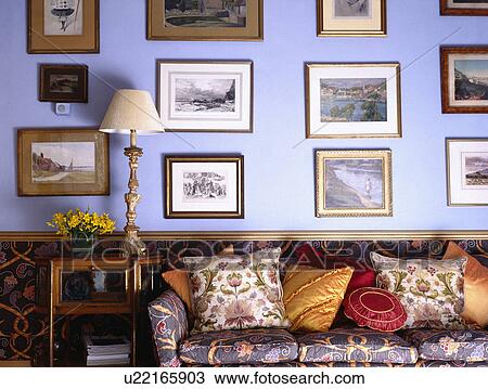 Stock Image - Group of pictures on blue living room wall above paterned sofa piled with colourful cushions. Fotosearch