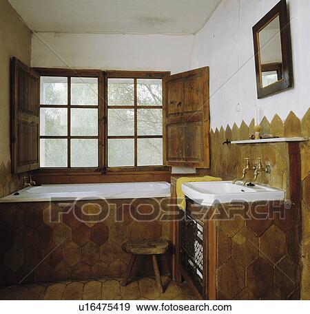 Stock Photo - Hexagonal clay tiles on bath and basin surrounds in Spanish bathroom with rustic wooden shutters on window above bath. Fotosearch