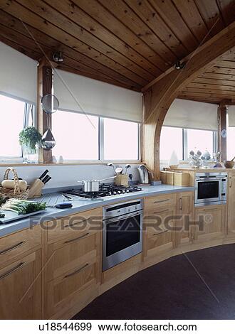 Stock Photo - Hob and oven in pale wood fitted cupboards below windows with white blinds in curved modern kitchen. Fotosearch