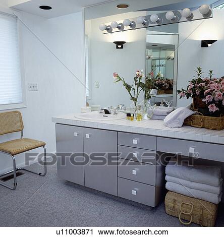 Stock Image - Hollywood-style lights on mirror above uplighters and basin in grey vanit unit with towels stored on lidded basket. Fotosearch