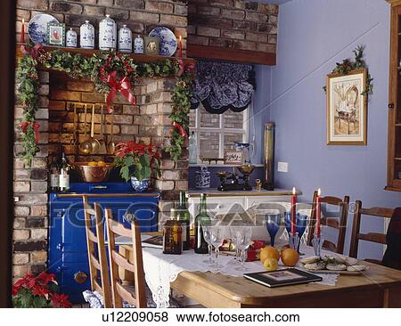 Stock Photo - Ivy garland above brick fireplace with blue Rayburn oven in blue country kitchen with table set for Christmas lunch. Fotosearch