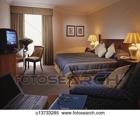 Laptop on table in foreground of hotel bedroom with lighted lamps on either side of bed and television on shelf View Large Photo Image Stock Photography - Laptop on table in foreground of hotel bedroom with lighted lamps on either side of bed and television on shelf. Fotosearch