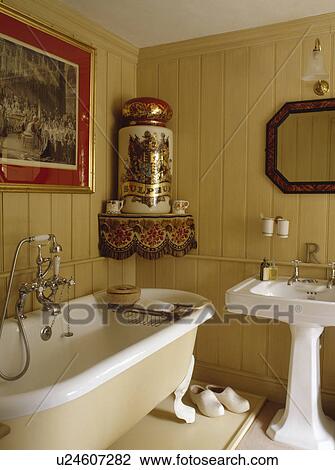 Stock Image - Large antique pot on corner shelf above clawfoot bath in neutral panelled bathroom with white pedestal basin. Fotosearch